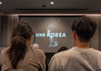 Two people sit facing a screen displaying the text "ONE KOREA" with a glowing map of the Korean Peninsula in a dimly lit room.