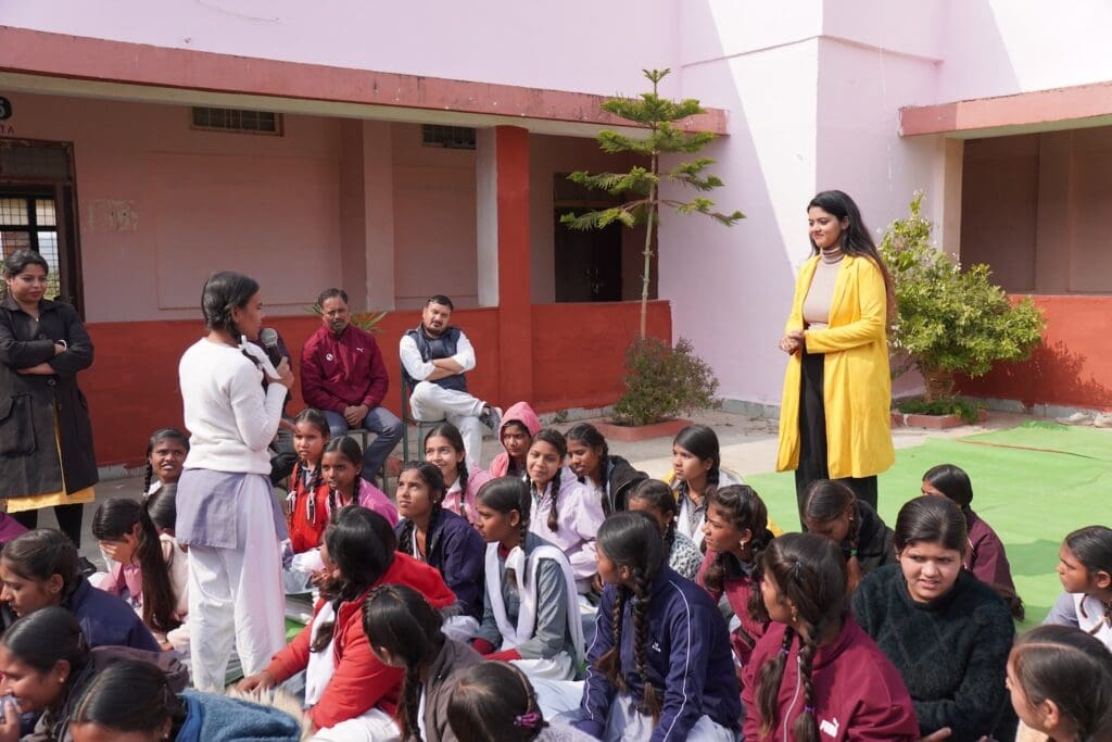 A group of schoolgirls sit outdoors while one stands to speak; a woman in a yellow coat listens attentively at the front, with other adults observing in the background during the Empowering Youth PeaceHub Campaign.