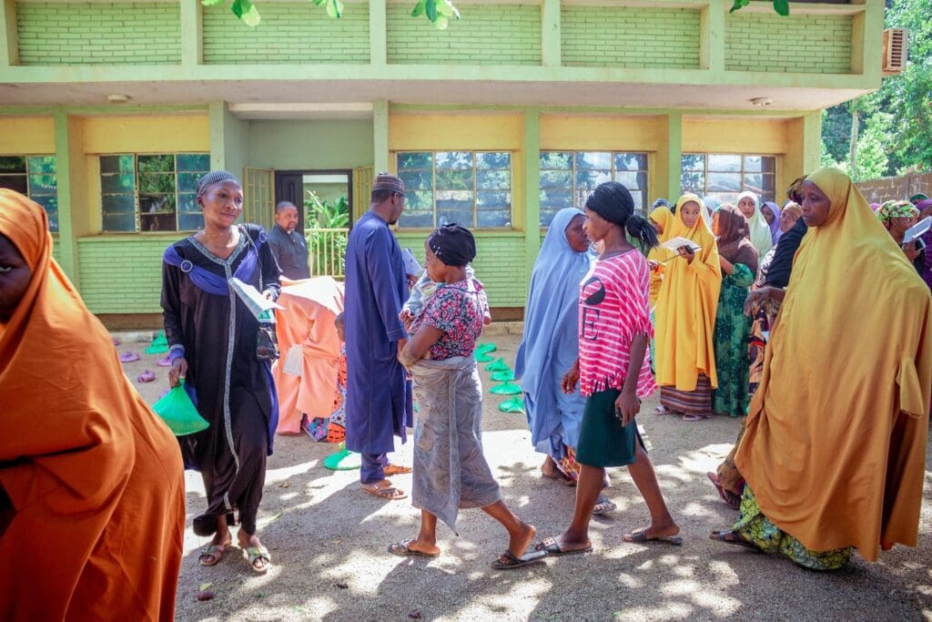 A group of women in Nigeria, mostly dressed in colorful clothing and headscarves, stand and walk outside a green building on a sunny day, reflecting interfaith harmony.