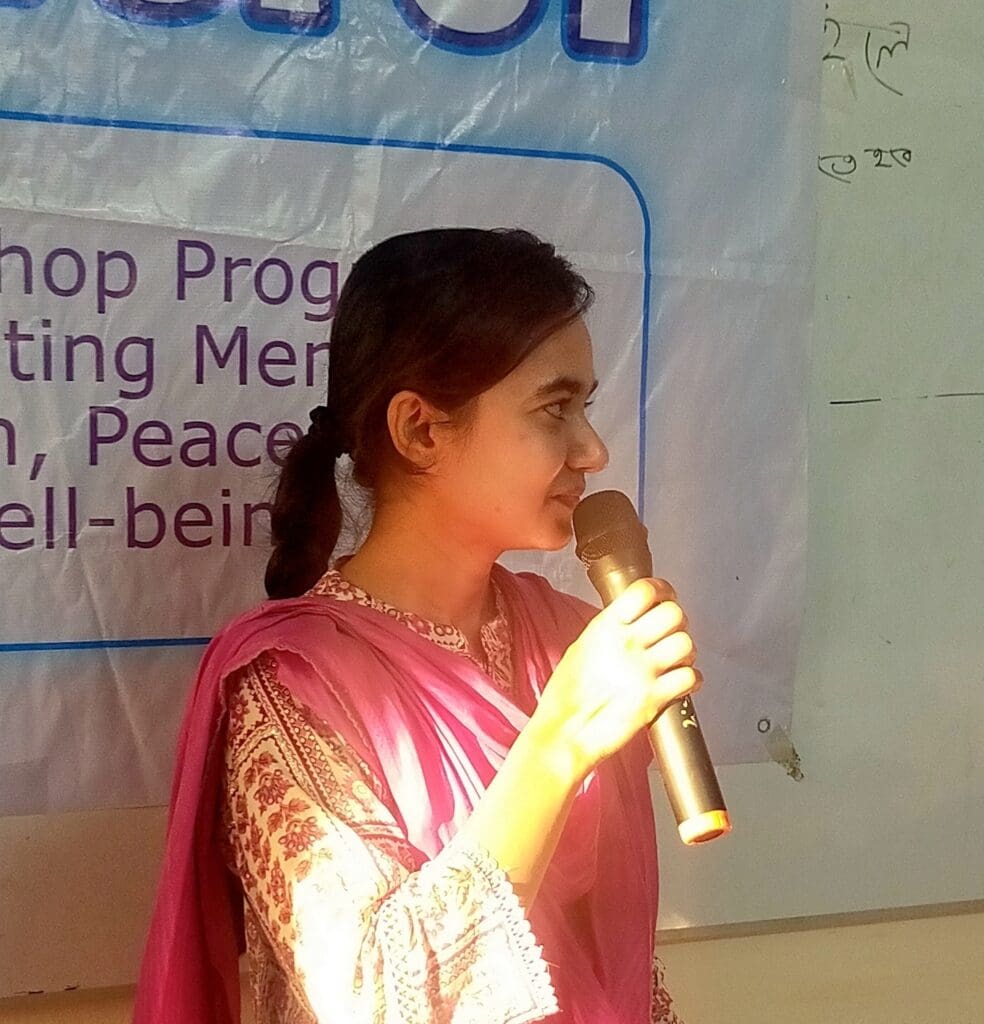 A woman in a pink outfit holds a microphone and speaks about well-being, standing in front of a whiteboard and banner during an event or workshop in Gazipur, Bangladesh.