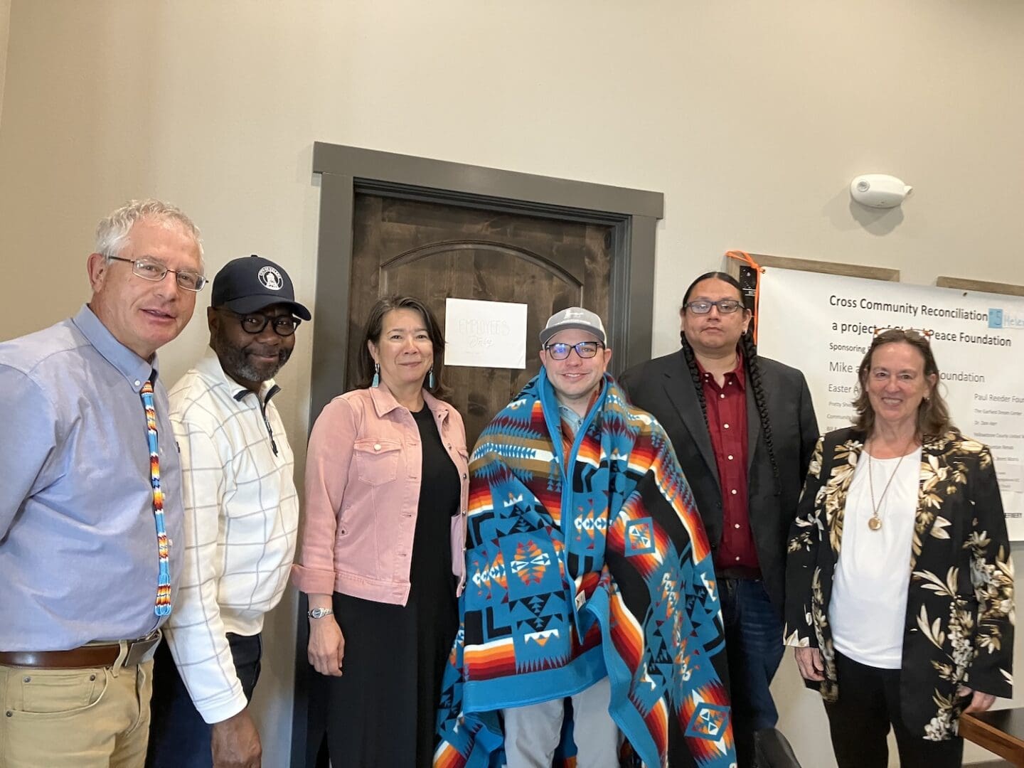 Six people stand indoors in front of a door and a sign, with one in the center wearing a patterned blanket. All are dressed in business or casual attire, gathered to support cross-community reconciliation and healing divides in Montana.