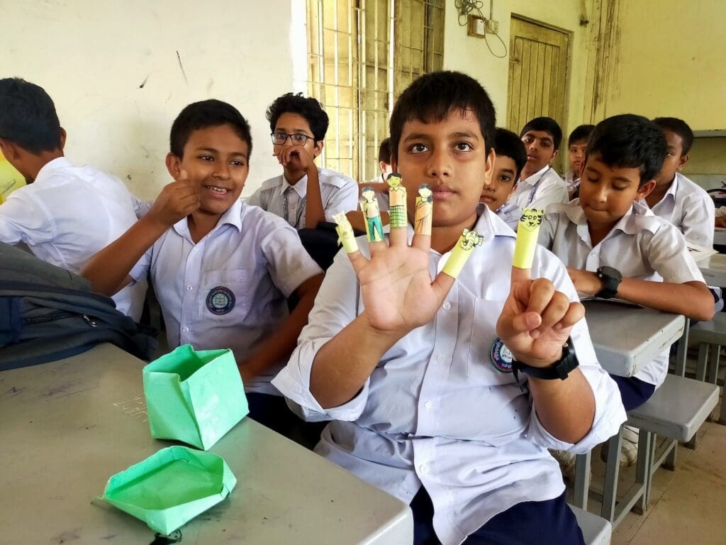 Boys in school uniforms sit at desks in a classroom in Gazipur, Bangladesh, with one boy in the foreground displaying finger puppets on his hand. There is a green paper box on the desk, fostering creativity and community well-being.