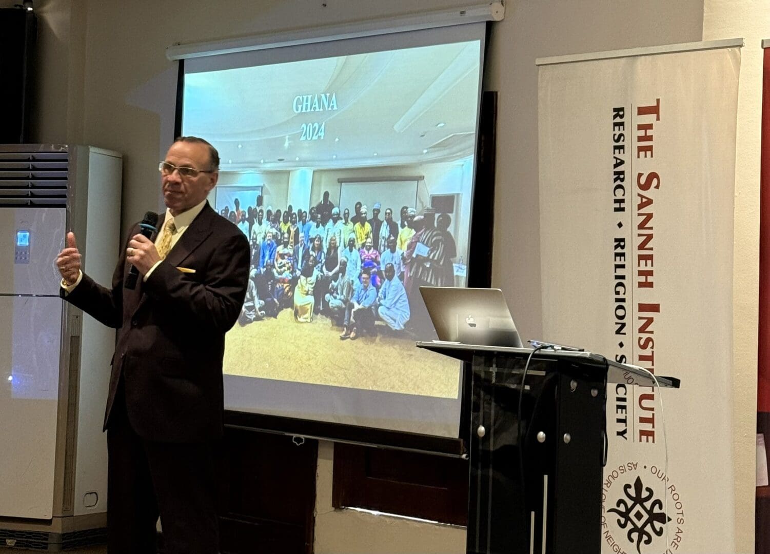 A man in a suit speaks at a podium with a laptop; a slide behind him reads
