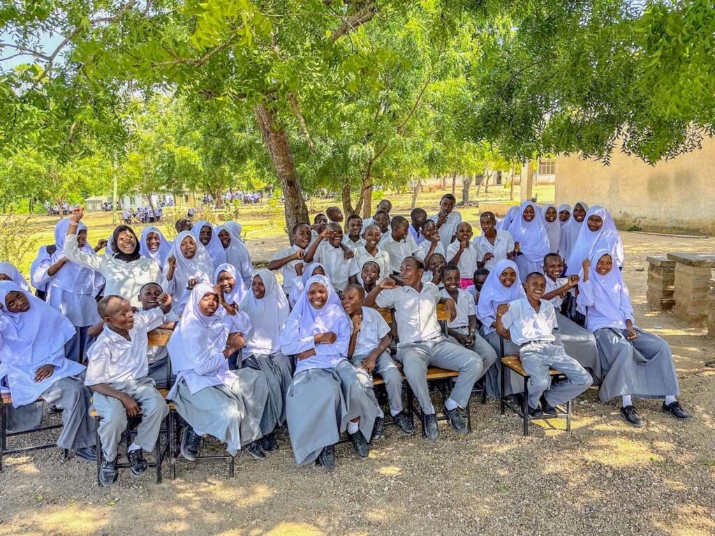 A group of students in school uniforms, including girls in white hijabs, sit and pose for a photo outdoors under a tree, showing unity and the hope for stronger families.