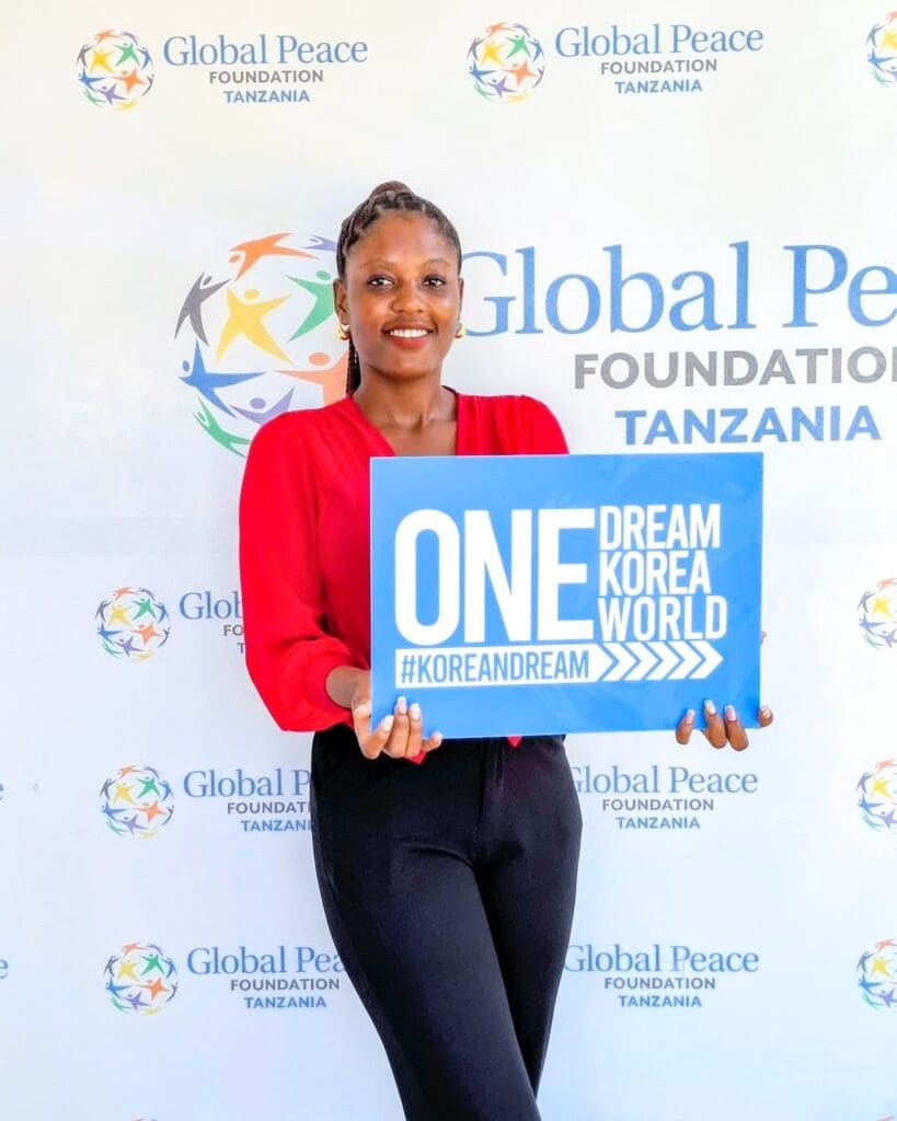 A woman stands in front of a Global Peace Foundation Tanzania backdrop, holding a sign that reads 