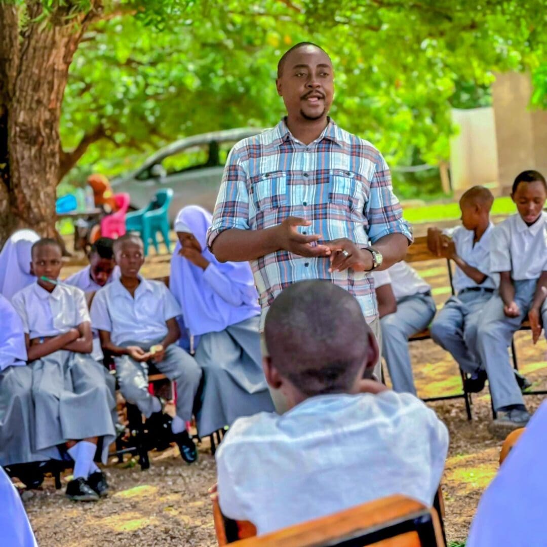 A man stands and speaks to a group of students seated outdoors in a circle under a tree, raising awareness about the impact of gender-based violence as the uniformed students listen attentively.