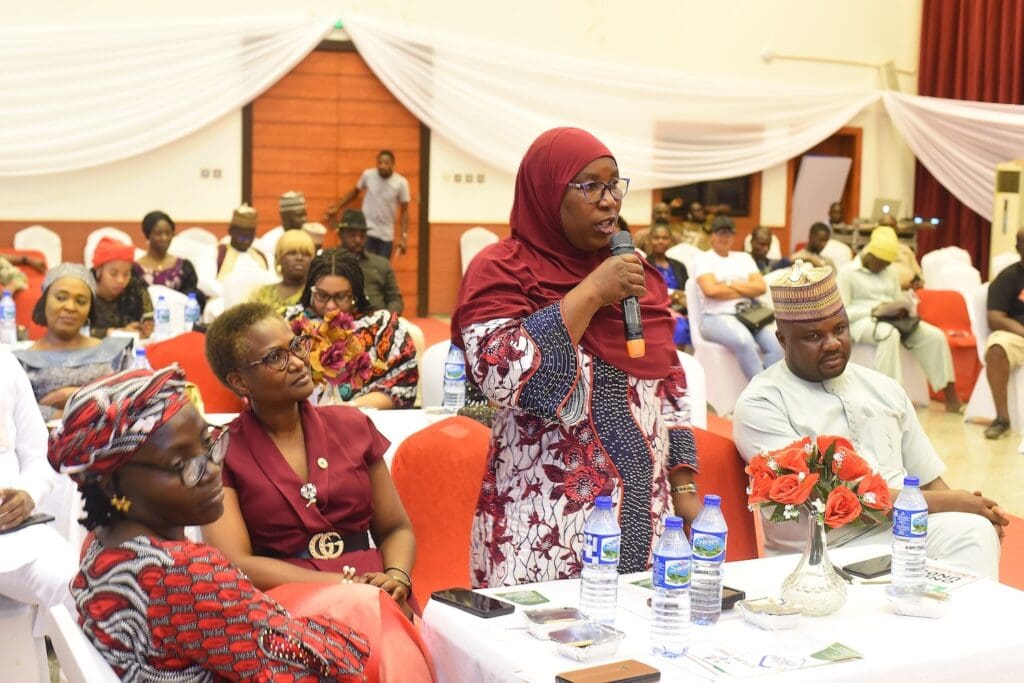 A woman in a maroon and white outfit stands with a microphone, speaking to an audience seated at tables in a decorated indoor event space.