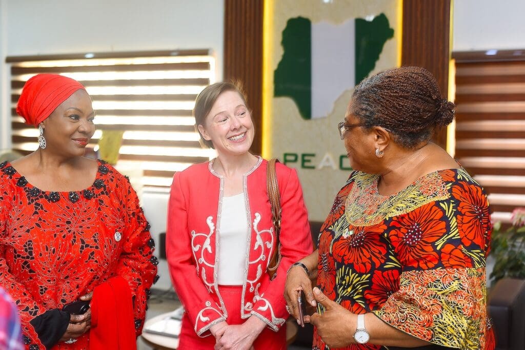 Three women stand together indoors, engaged in conversation; a map of Nigeria and the word 