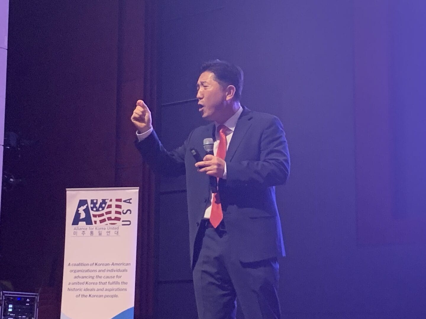 A man in a suit and red tie speaks into a microphone on stage next to a banner that reads