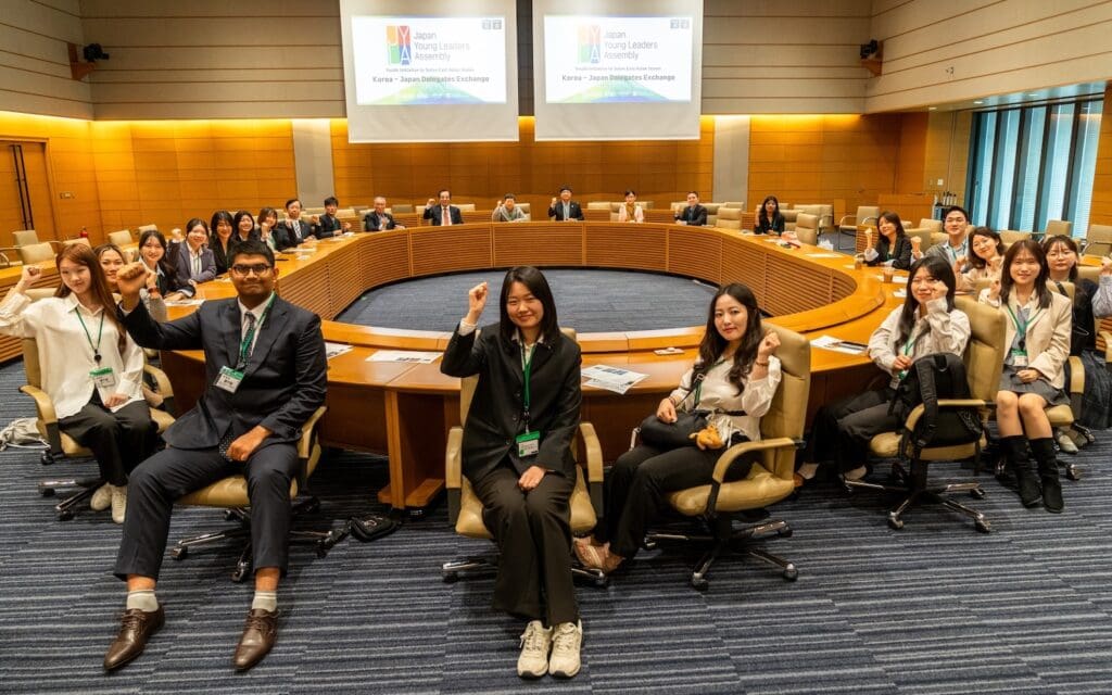 A group of youth leaders sit around a large circular conference table in a modern meeting room, with presentation slides about peace on the Korean Peninsula displayed on two screens at the front.
