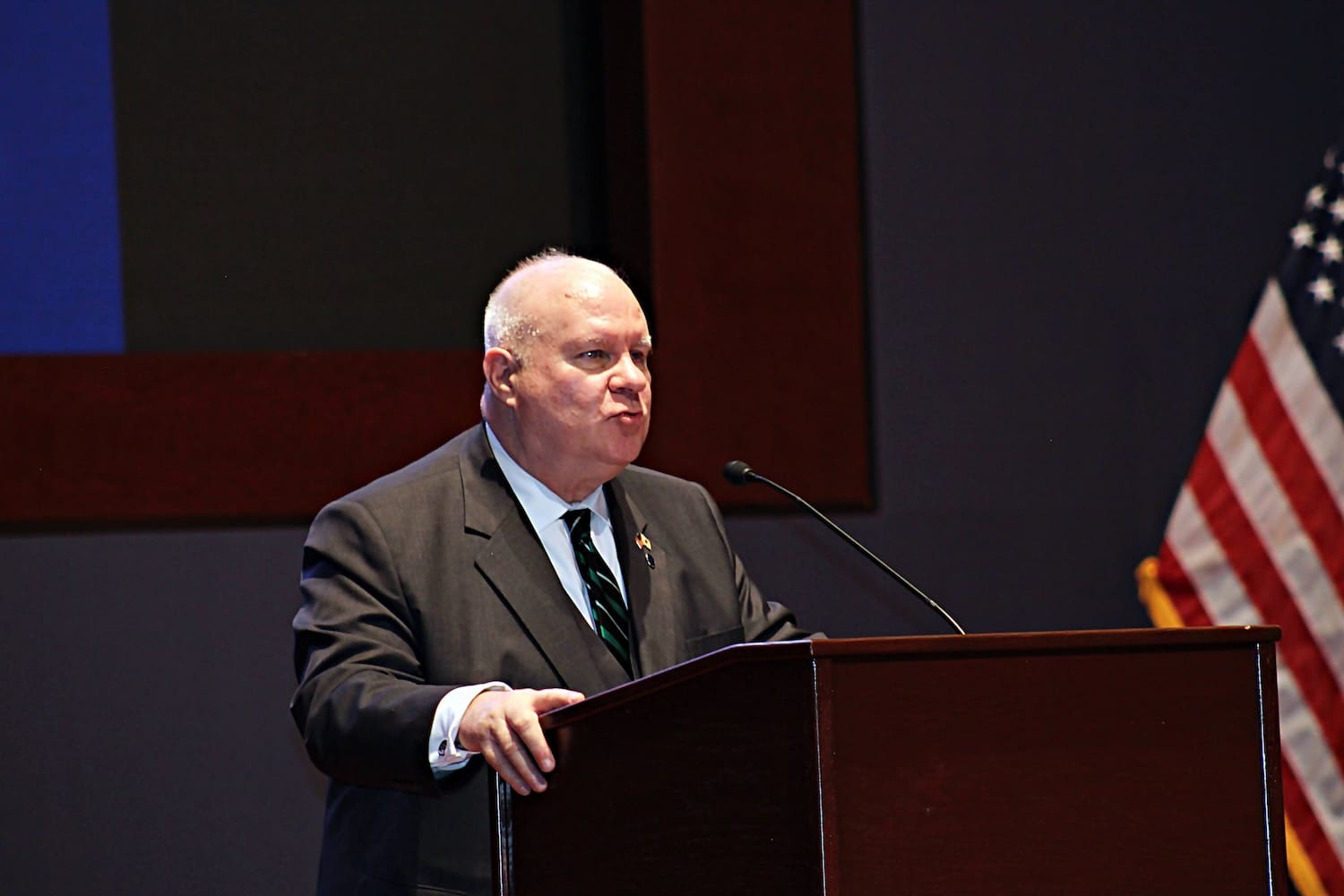 A man in a suit speaks at a podium with a microphone during the Capitol Forum; an American flag is visible in the background.