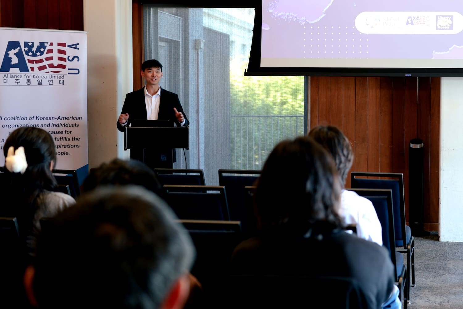 A man gives a presentation at a podium to an audience in a conference room, with a USA-Korea alliance banner and a projection screen, highlighting the importance of youth leadership for a unified Korea.