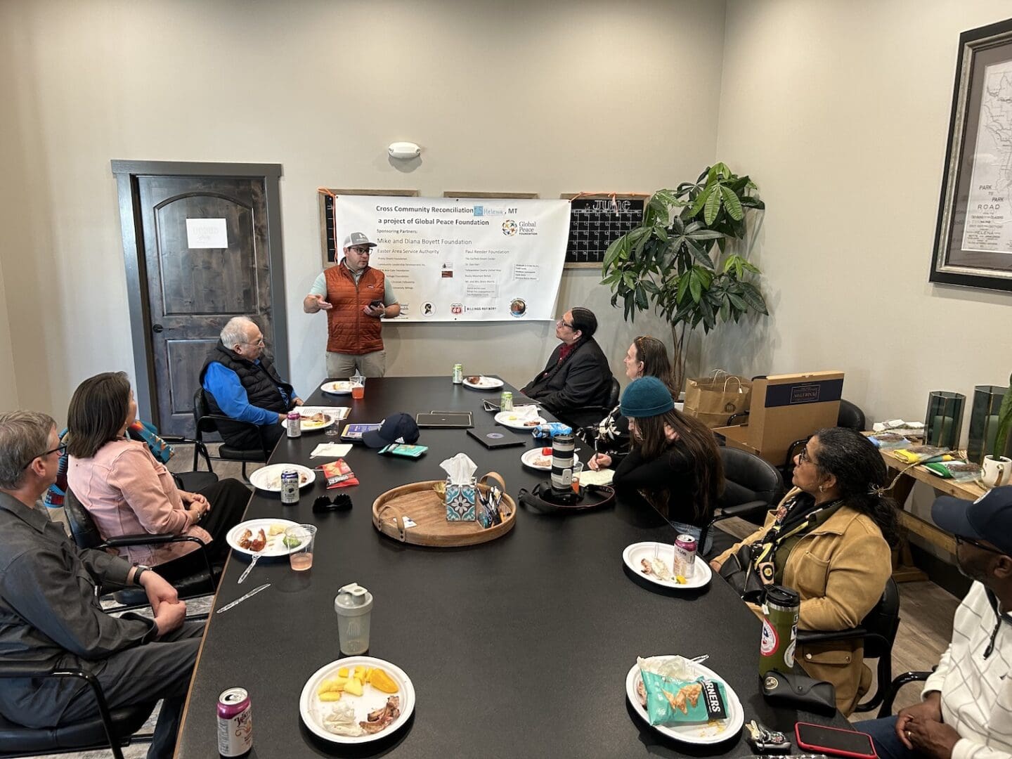 A woman stands and speaks to a group of people seated around a conference table in Montana, with a presentation board displayed behind her and food and drinks on the table, fostering cross-community reconciliation.