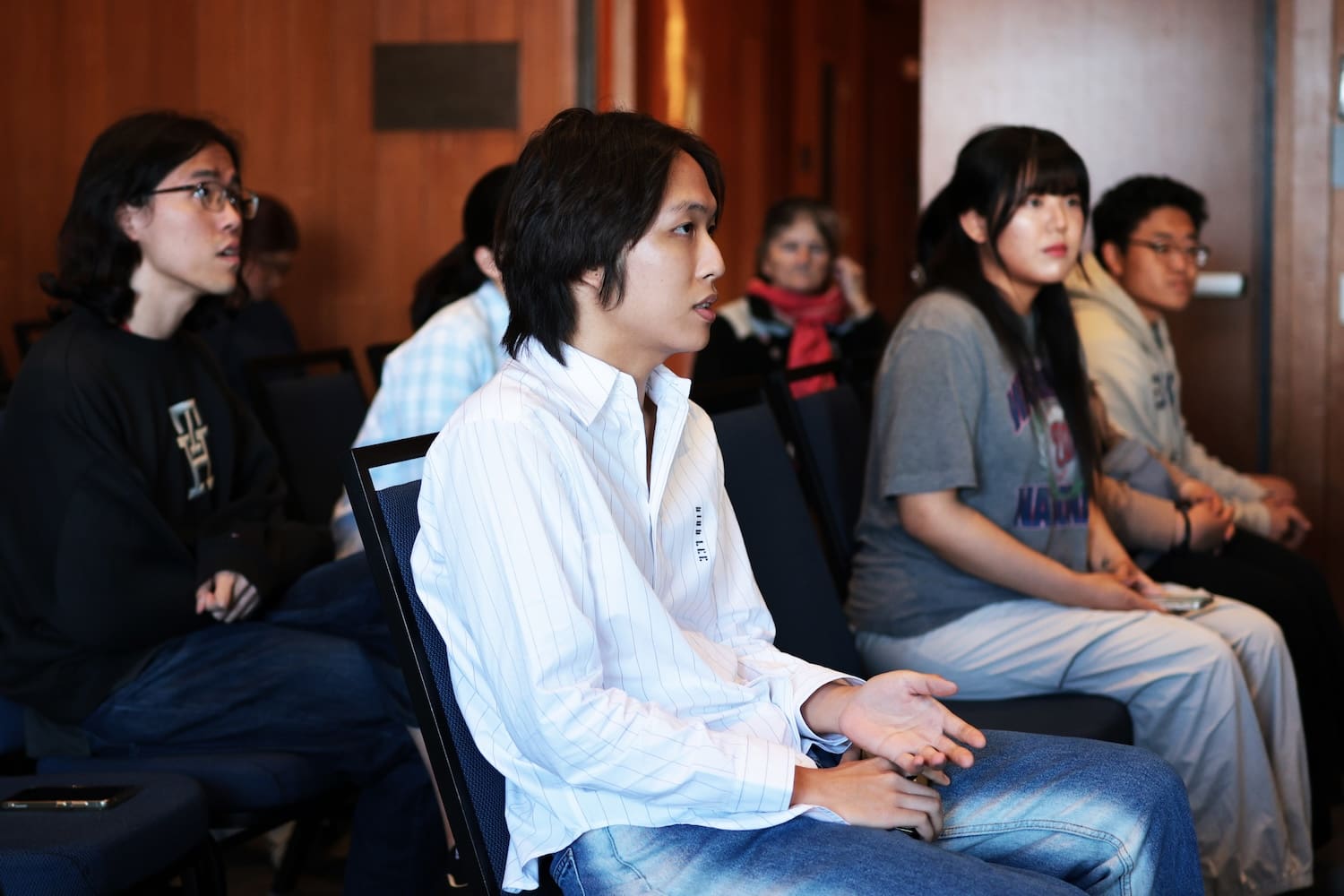 A group of people sit indoors in rows of chairs, listening attentively; one person in the foreground gestures with their hand while speaking about youth leadership and the vision of a unified Korea.