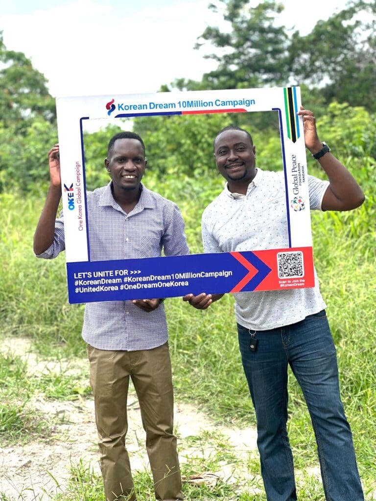 Two men standing outdoors in grass, holding a large rectangular frame promoting the Korean Dream 10 Million Campaign and celebrating Global Unity.