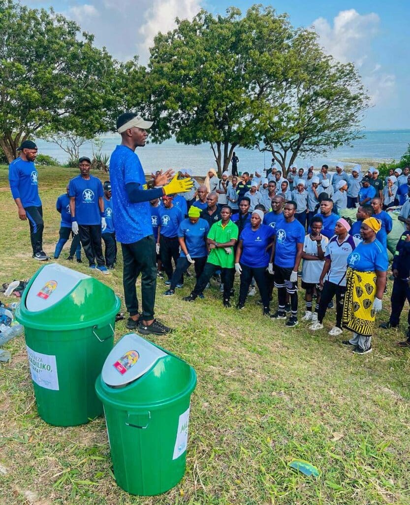 A man addresses a group of people in blue shirts near two green recycling bins outdoors, with trees and a body of water behind them, highlighting community power in tackling coastal pollution.