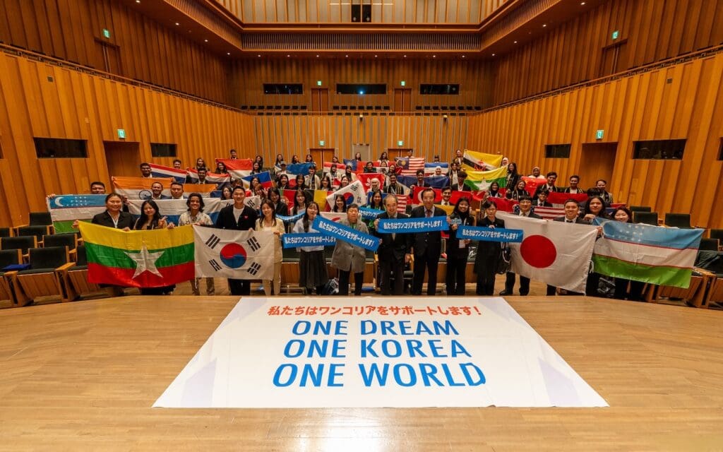 A diverse group of people stands in an auditorium holding various national flags behind a large banner that reads