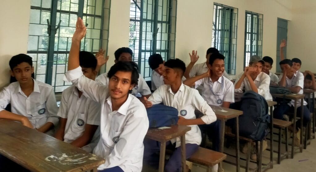 A group of male students in school uniforms sit in a classroom in Gazipur, with several raising their hands and others looking forward or talking, reflecting their engagement and well-being.