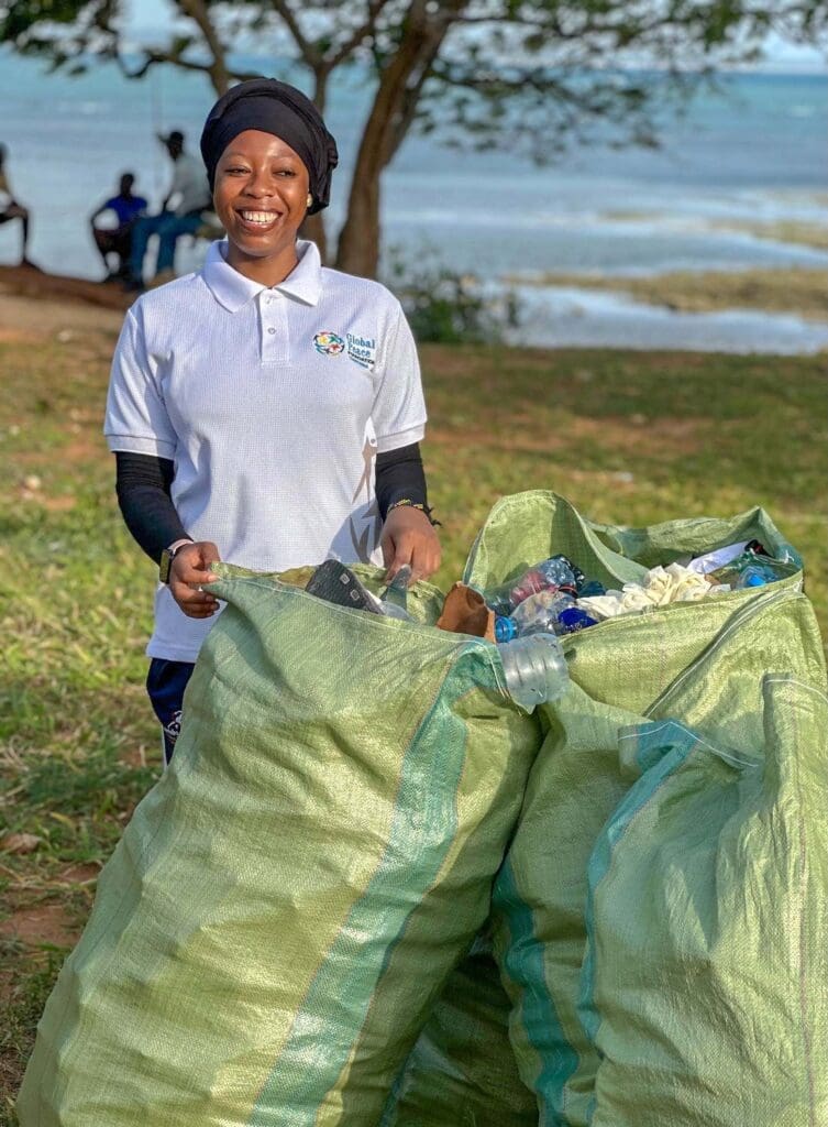 A woman in a white polo shirt stands outdoors in Tanzania, smiling beside two large green bags filled with collected plastic waste—a symbol of community power against coastal pollution.