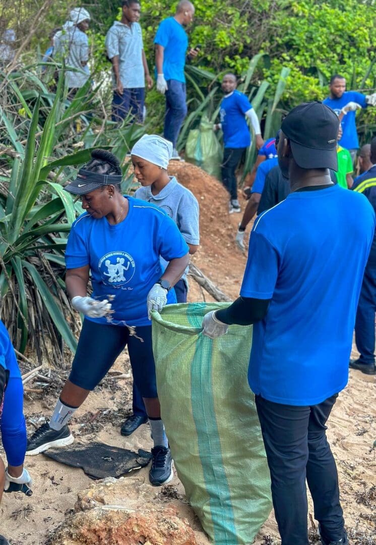 A group of people in blue shirts and gloves harness community power in Tanzania, cleaning up a natural outdoor area by collecting coastal pollution and placing debris into large green bags.