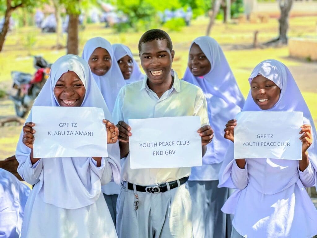 A group of students in uniforms hold signs promoting peace clubs and ending gender-based violence, smiling outdoors in daylight as they advocate for stronger families.