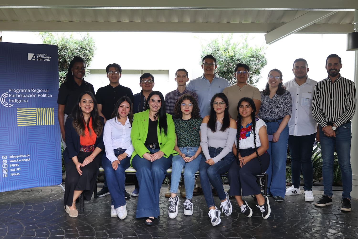 A group of fourteen people, including Indigenous Youth Leaders, pose indoors—some seated, others standing—before a banner for a regional indigenous political participation program.