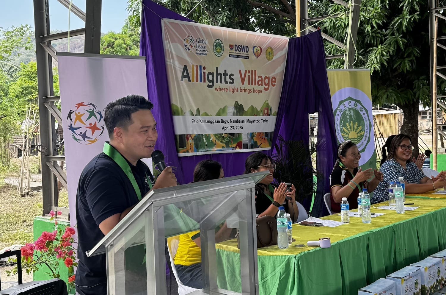 A man speaks at a podium during the All-Lights Village Program event, focused on sustainable development, with several people seated at a table on stage and banners and decorations in the background.