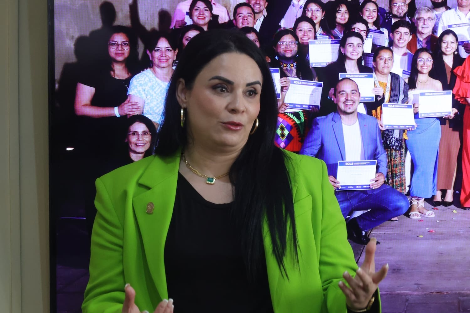 A woman in a bright green blazer speaks in front of a group photo display featuring Indigenous Youth Leaders from Central America, holding certificates and posing proudly for the camera.
