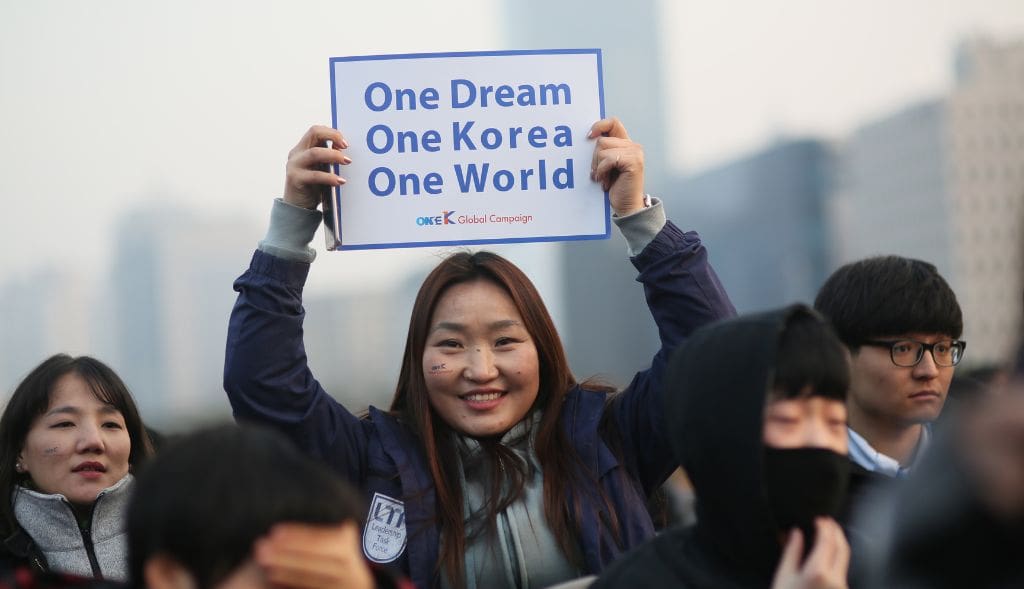 A woman in a crowd holds up a sign that reads, “One Dream One Korea One World,” symbolizing the spirit of One Korea Membership. Other people stand around her, some wearing masks and winter clothing.