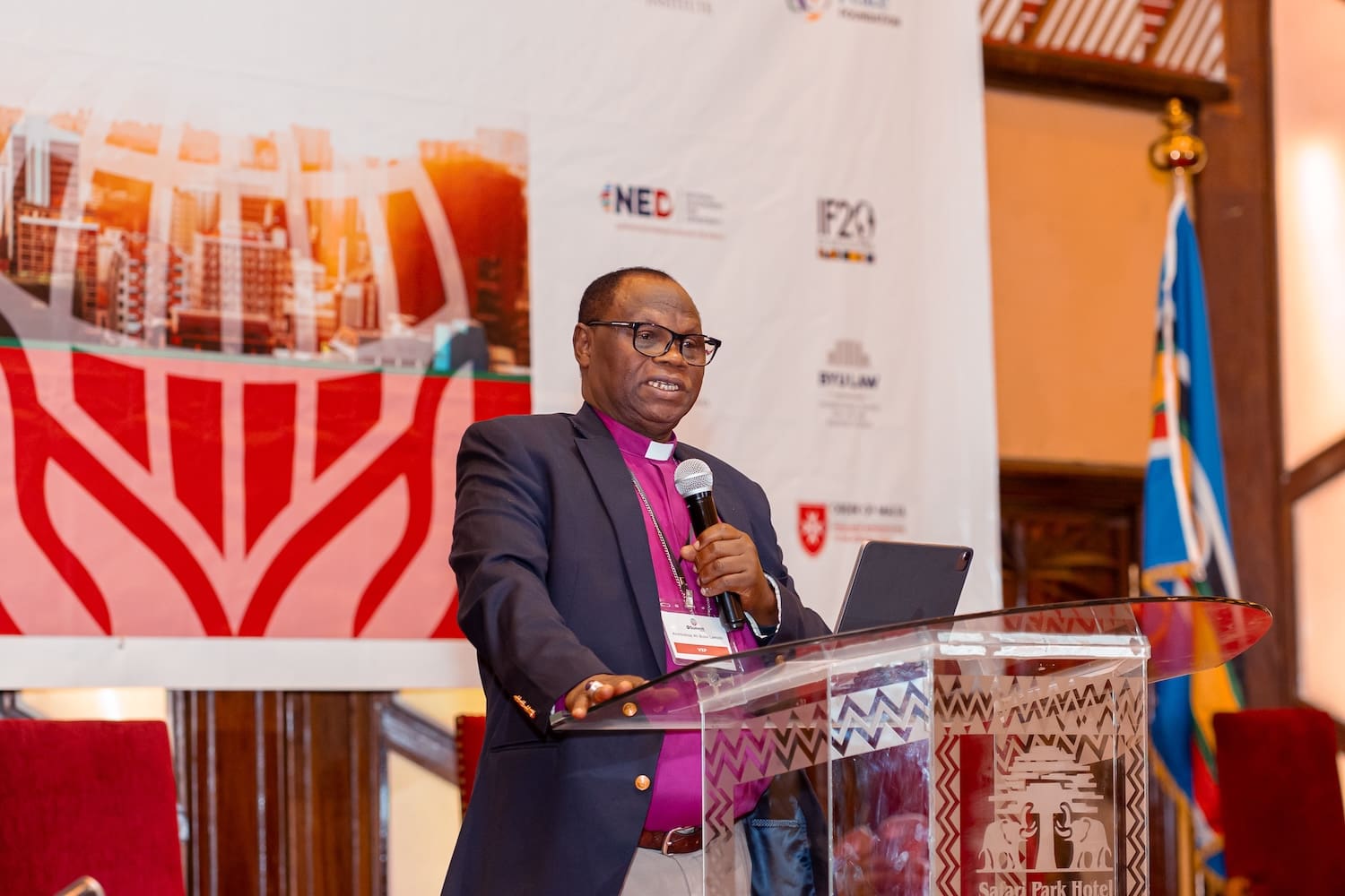 A man in a suit and clerical collar speaks at a podium during the IRF Summit Africa, with a microphone, laptop, and event banner in the background.