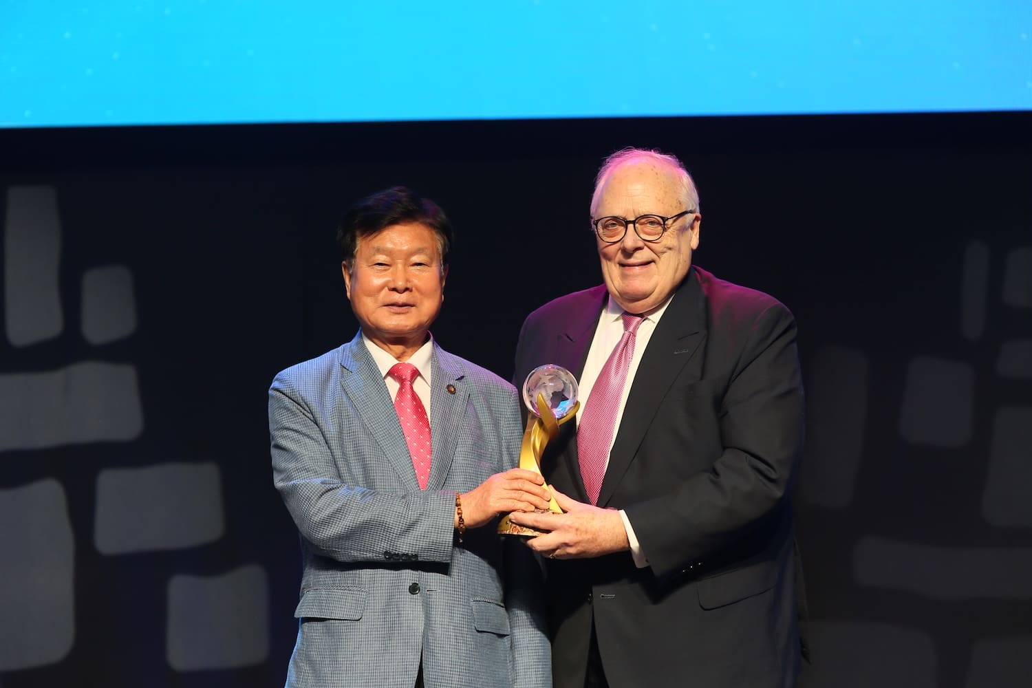 Two men in suits stand on stage, one handing an award trophy to Dr. Edwin J. Feulner, both facing the camera and smiling.