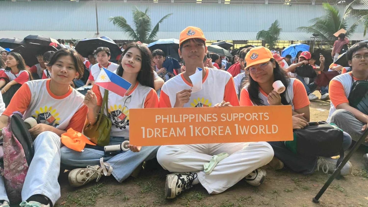 A group of young people wearing matching shirts and caps sit together holding a sign that reads,