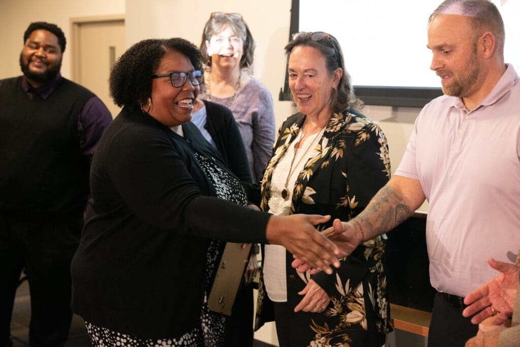 A group of five adults interact indoors; a woman in black smiles and extends her hand in a gesture of peace to a man in a light purple shirt during a Global Peace Foundation event in Baltimore.