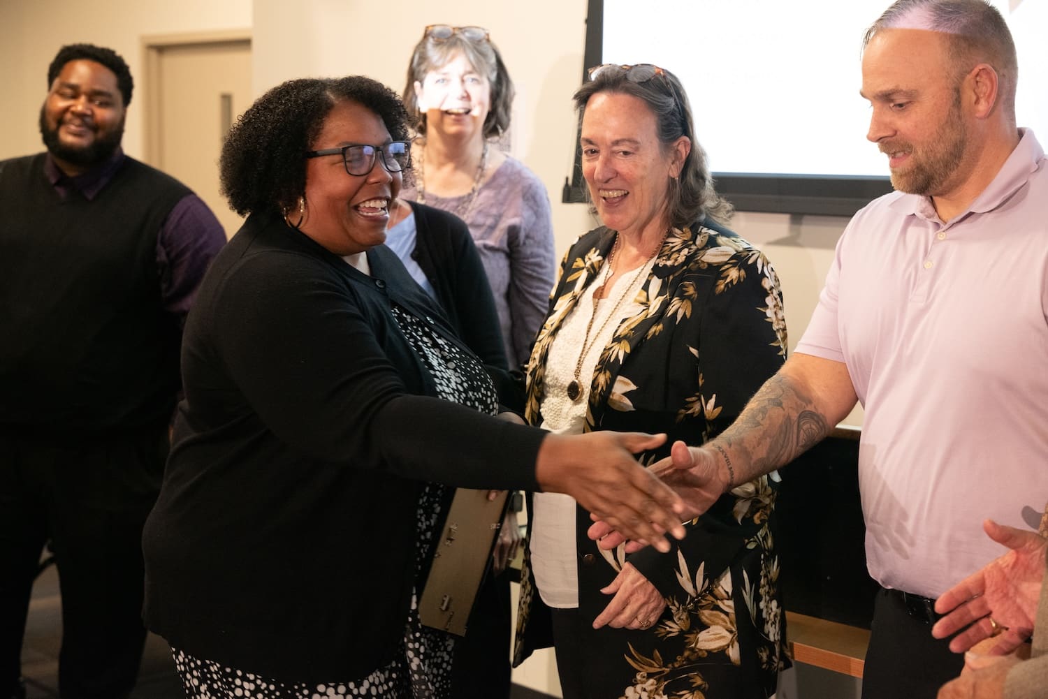 A group of five adults interact indoors; a woman in black smiles and extends her hand in a gesture of peace to a man in a light purple shirt during a Global Peace Foundation event in Baltimore.