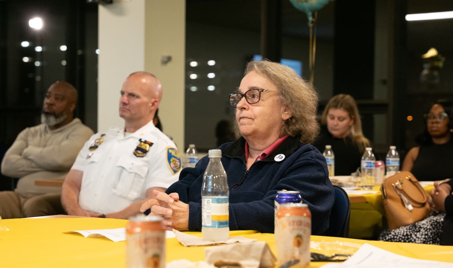 A group of people, including a uniformed officer, sit at a table with drinks and papers, attentively listening during an indoor reconciliation event.