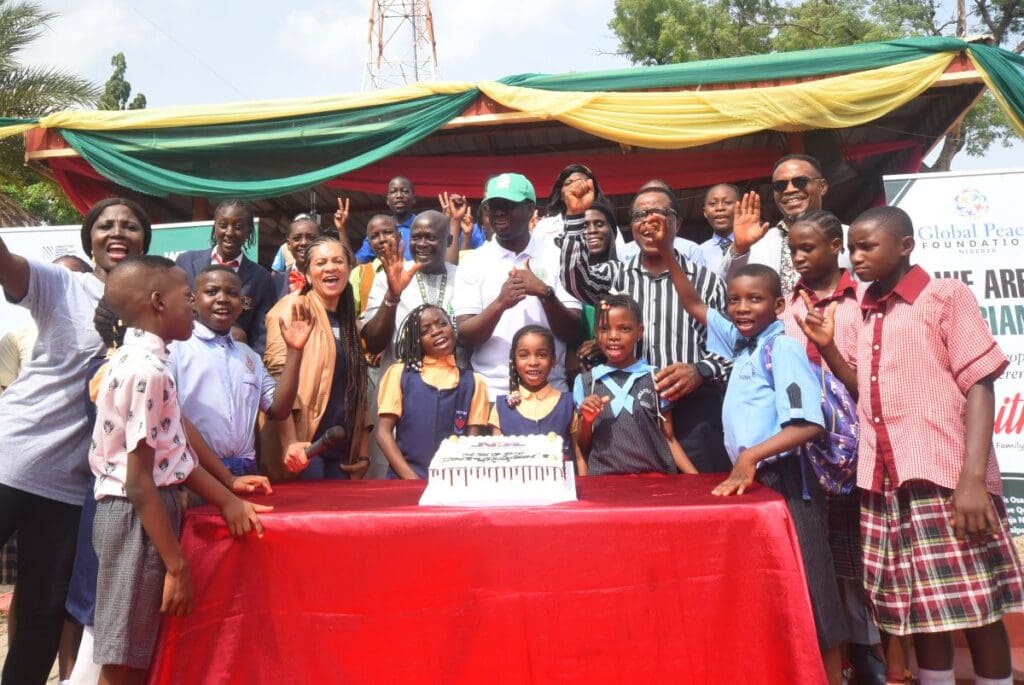 A group of adults and children pose around a cake outdoors, smiling for a photo at a GPF Nigeria event with banners and a decorated canopy in the background, celebrating efforts to empower children.