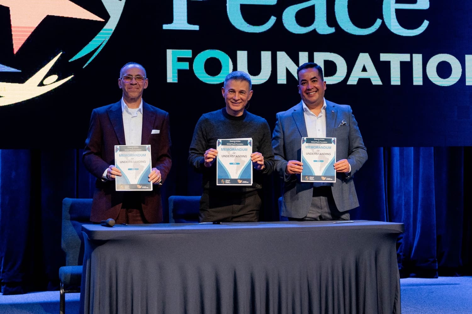 Three men in suits stand behind a table, each holding a certificate, with a large 
