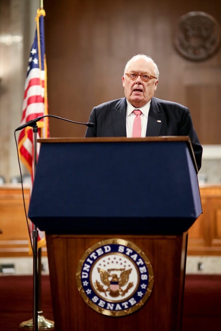 An older man in a suit and tie speaks at a podium with the United States Senate seal, honoring Dr. Edwin J. Feulner, with an American flag and official emblem visible in the background.