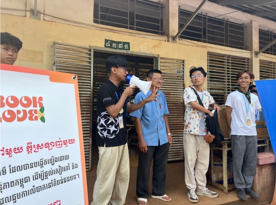 A group of people stand outside a building; one person from the Global Peace Foundation Cambodia speaks into a megaphone next to a sign with text in Khmer and English that reads