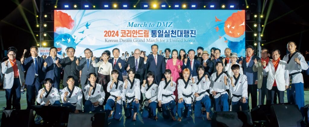 A large group of people, including officials and performers in uniforms, pose together onstage in front of a 
