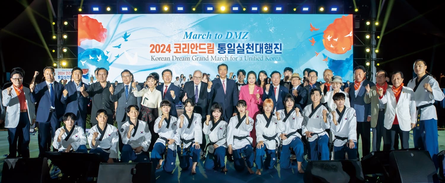 A large group of people, including officials and performers in uniforms, pose together onstage in front of a "March to DMZ" and "Korean Dream Grand March for a United Korea" event banner with Dr. Edwin J. Feulner present.
