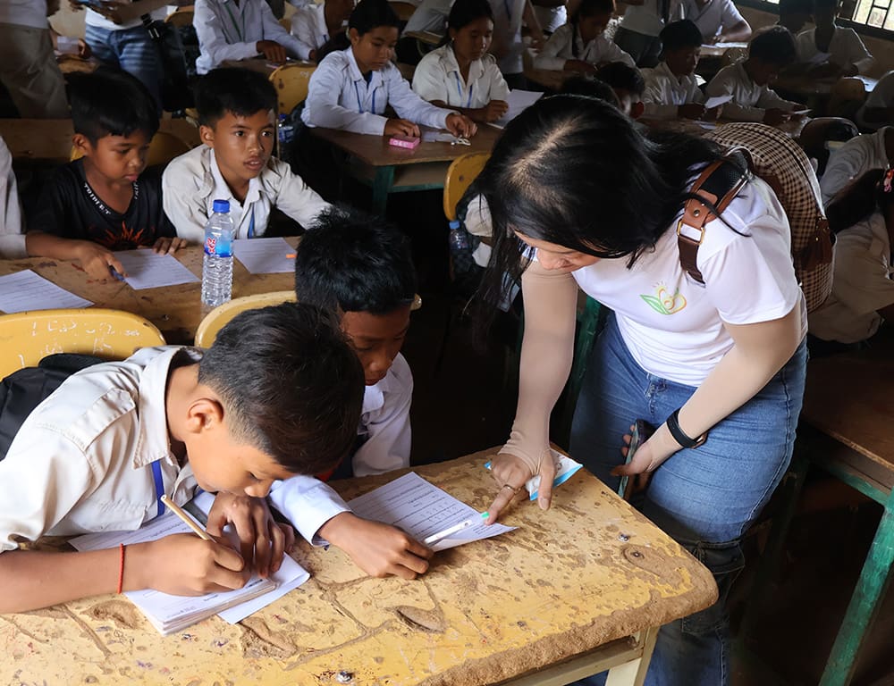 A teacher assists two students with their worksheets in a crowded classroom on International Youth Day 2025, while other children work at their desks in the background.