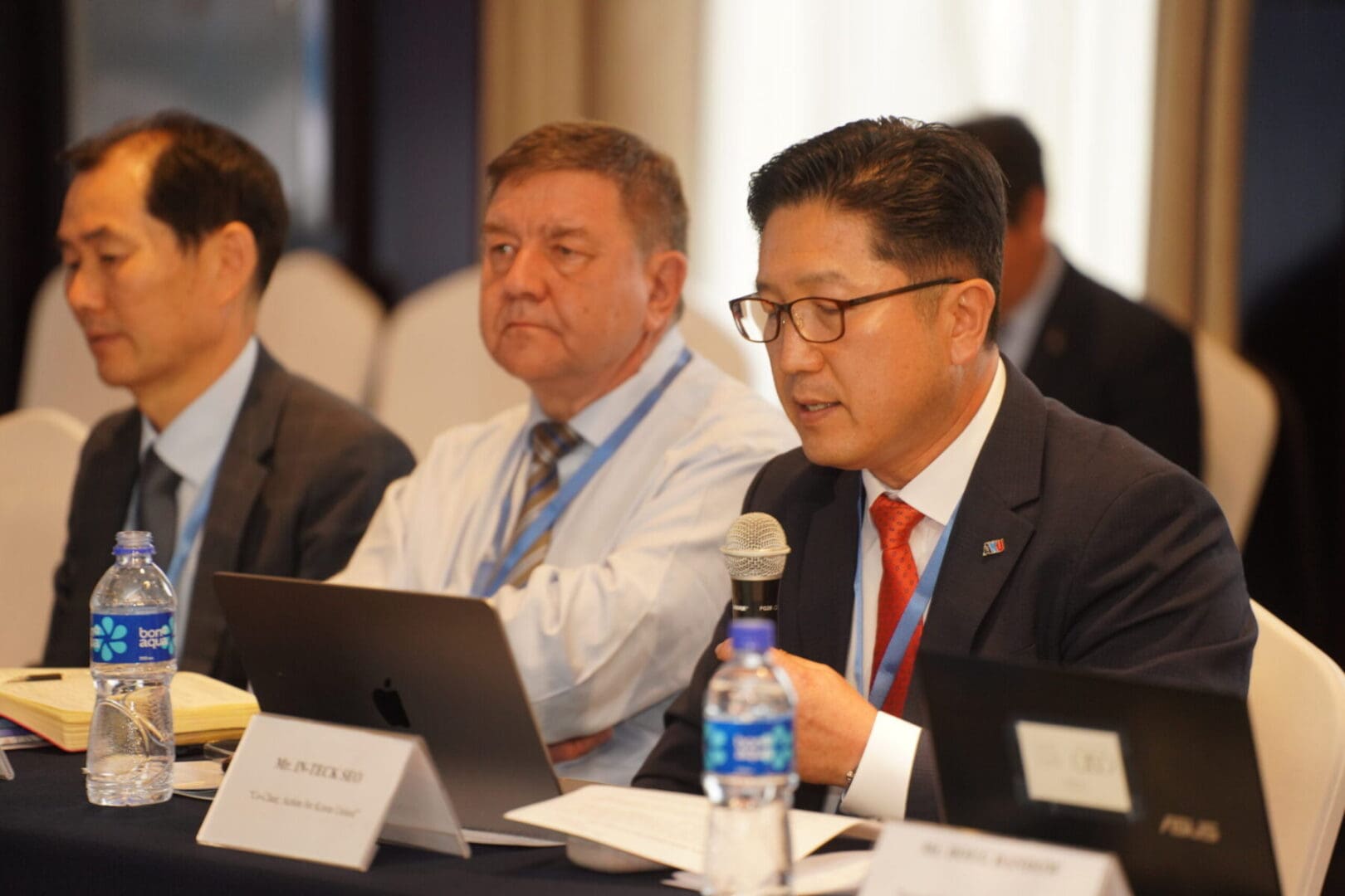 Three men in business attire sit at a conference table with laptops and water bottles; one man speaks into a microphone, addressing key issues such as Korean Unification and Mongolia’s role in Climate Action.