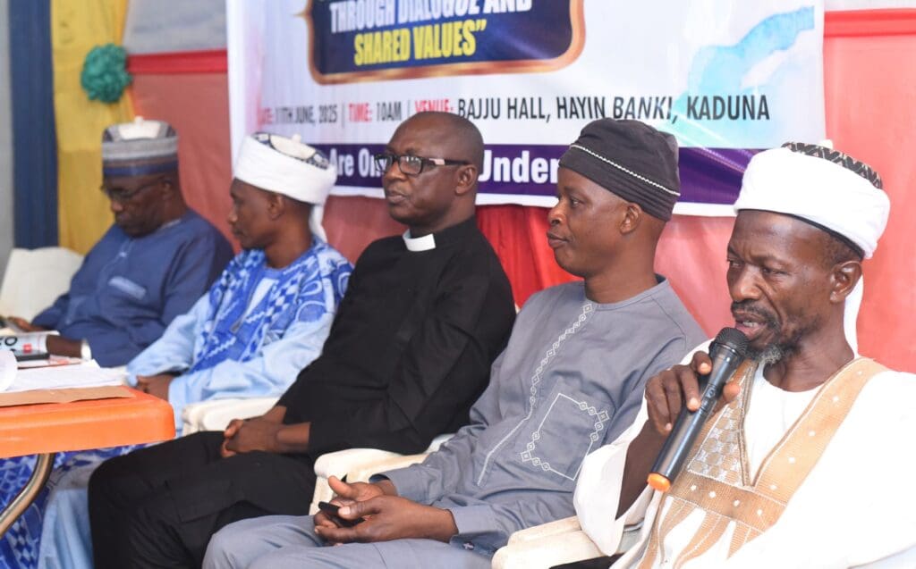 Five men, including religious leaders in traditional and clerical attire, sit in a row at an indoor event with a banner in the background. One man is speaking into a microphone, fostering dialogue and mutual respect among attendees.
