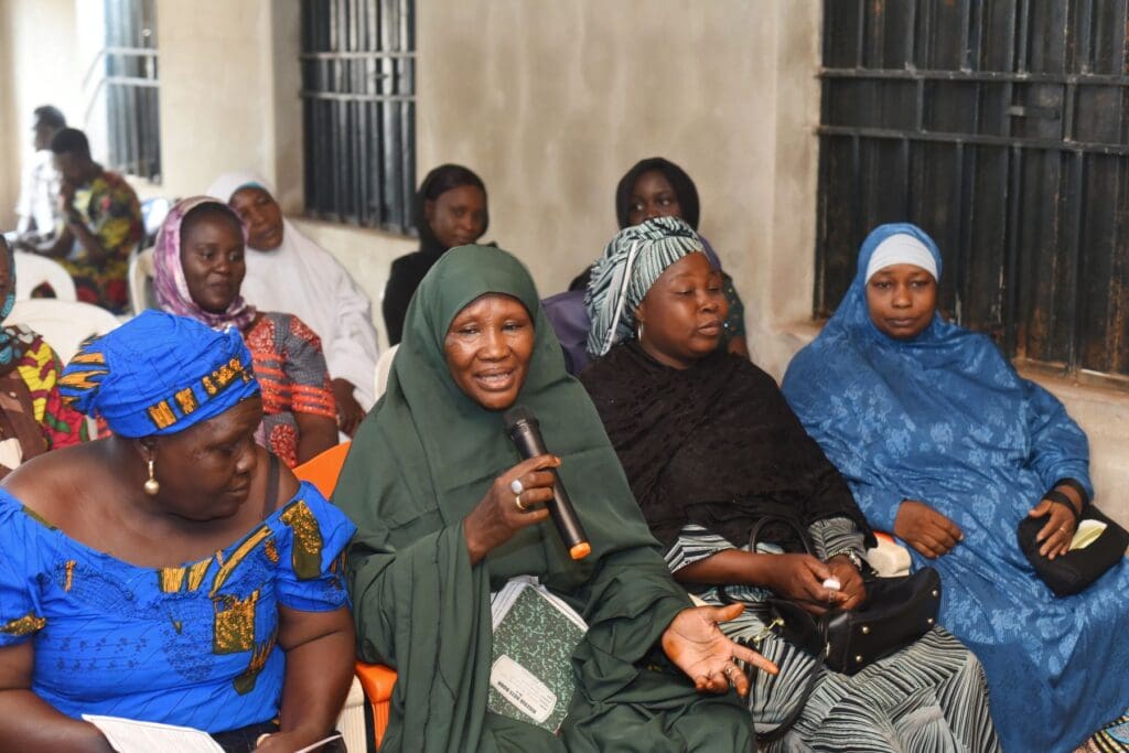 A group of women sit indoors; one woman in the center holds a microphone and speaks, fostering dialogue while others listen attentively, reflecting mutual respect.