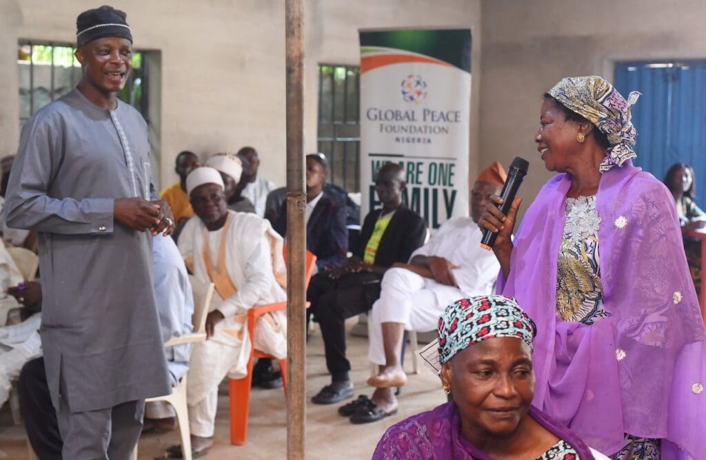 A woman in purple traditional attire speaks into a microphone while a man in grey stands nearby; others sit and watch in a gathering focused on dialogue and shared values, with a Global Peace Foundation banner in the background.