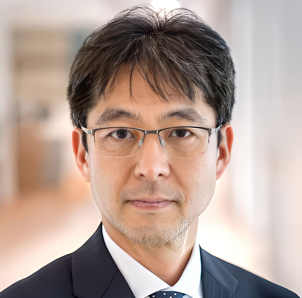 Komei Isozaki A middle-aged man with short dark hair, glasses, and a suit looks directly at the camera in a brightly lit indoor setting.