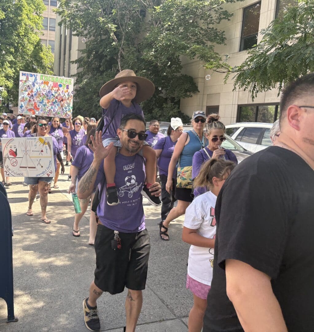 A group of people, many in purple shirts, walk outside in the 2025 Billings March Against Drugs parade. A man carries a child on his shoulders while others hold colorful signs. Trees and a building are in the background.