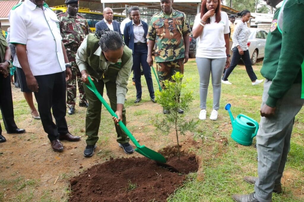 A woman uses a green shovel to plant a young tree from the tree nursery at Wareng High School, with several people from GPF Kenya and others in uniforms and casual attire standing nearby.