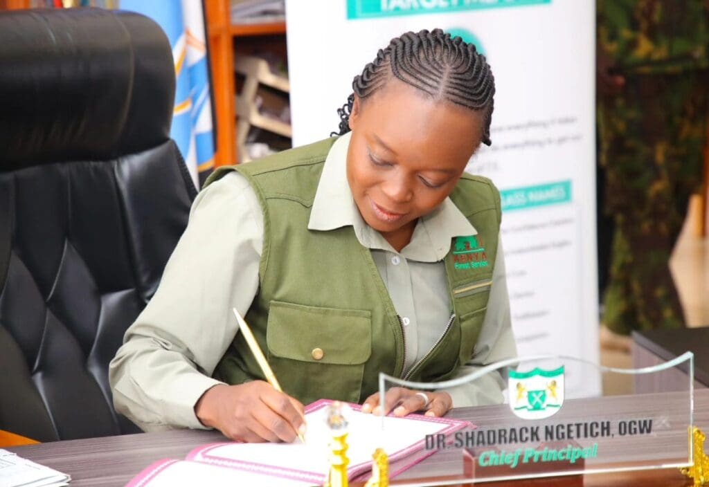A woman in uniform sits at a desk, writing on paper with a pencil. A nameplate reading